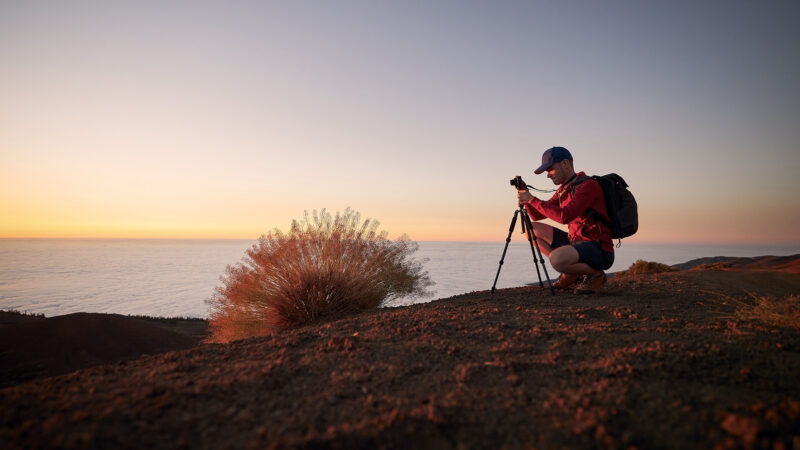 Young man during photographing landscape on top of hill above clouds. Hobby photographer waiting for beautiful sunset. Tenerife, Canary Islands, Spain.